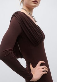 Καφέ φόρεμα με μακριά μανίκια, ντεκολτέ με draping και εφαρμοστή σιλουέτα, με ομαλή υφή και άνοιγμα στο πλάι. Διακοσμημέν�ο με ασημένιο κολιέ αλυσίδα.