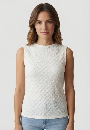 Femme aux cheveux bruns ondulés, portant un haut blanc texturé sans manches et un jean bleu clair, sur un fond clair uni.