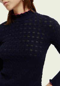 Granatowy dzianinowy sweter z teksturowanym wzorem w romby. Posiada kołnierz typu stójka z różowym falbankowym wykończeniem oraz dopasowane rękawy.