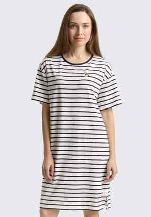 Vrouw die een witte knie-lange jurk draagt met zwarte horizontale strepen en een kleine geborduurde bij op de borst, staand tegen een effen achtergrond.