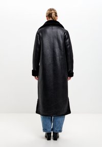 Dlouhý černý kabát s měkkým límcem, texturovanou tkaninou a kontrastními manžetami. Obsahuje boční zipy a rovný střih, kombinovaný s modrými džíny.