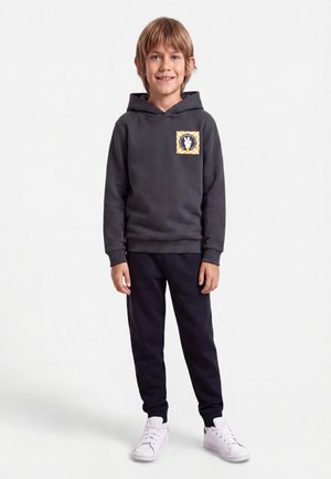 Garçon portant un sweat à capuche gris foncé avec un patch graphique, un pantalon noir et des baskets blanches, debout et souriant devant un fond uni.