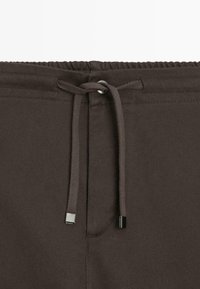 Ciemnobrązowe spodnie dresowe z elastycznym pasem, sznurkiem do regulacji i srebrnymi ozdobami. Posiadają ukośną fakturę.