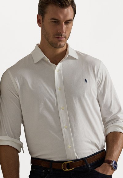 Λευκό πουκάμισο με κουμπιά, φτιαγμένο από μαλακό υλικό, με κλασικό γιακά, γυρισμένα μανίκια και ένα μικρό μπλε έμβλημα polo στο στήθος.