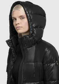 Black puffer jacket with a shiny finish, featuring a detachable hood, button closure, and drawstring accents on the collar.