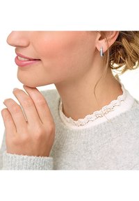 Orecchini a cerchio in argento con una finitura testurizzata, indossati da una persona contro un morbido maglione grigio con dettagli in pizzo. Mano che tocca delicatamente il viso.