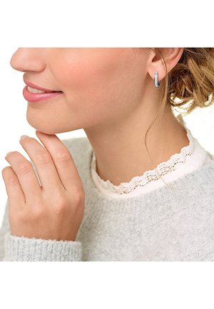 Silberne Creolen mit strukturiertem Finish, getragen von einer Person mit einem weichen, grauen Pullover mit Spitzenakzenten. Hand berührt sanft das Gesicht.