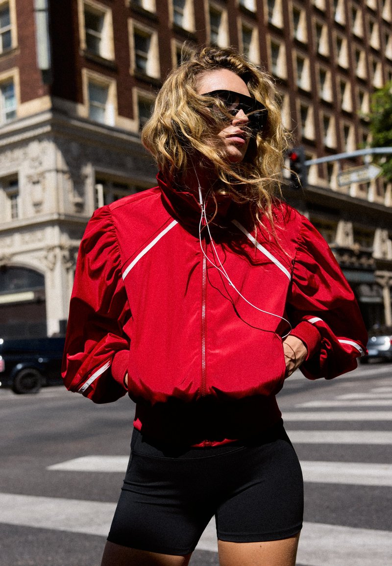 Femme aux cheveux blonds bouclés, veste rouge et short noir, debout dans une rue de la ville, portant des lunettes de soleil et des écouteurs.