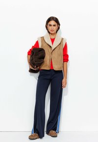 Gilet en fausse fourrure marron avec doublure crème, pull rouge, pantalon large navy avec rayure blanche et chaussures à imprimé léopard marron.