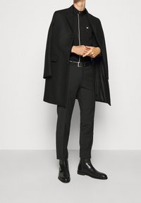 Cappotto nero indossato sopra una camicia nera con bottoni e piping bianco, pantaloni neri e stivali alla caviglia neri. Linee pulite e vestibilità su misura.