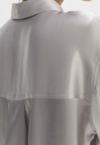 Chemise satin gris clair avec un col classique, texture lisse et une couture au dos. Design simple et élégant.
