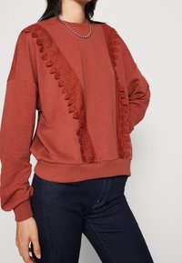 Terakotová mikina s krajkovými detaily na ramenou, dlouhými rukávy a žebrovanými lemy. Kombinováno s tmavými džínami pro ležérní vzhled.
