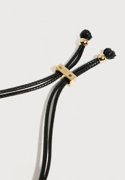 Ρυθμιζόμενο μαύρο πλεγμένο κορδόνι με κούμπωμα ολίσθησης σε χρυσό τόνο και τελικά καπάκια σε απλό ανοιχτό φόντο.