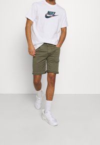 Vit bomull t-shirt med en flerfärgad Nike-logotyp, kombinerad med olivgröna shorts och vita sneakers. Shortsen är uppvikta vid kanten.