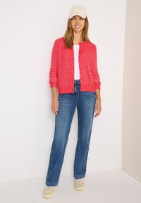 Cardigan rouge texturé avec des boutons, t-shirt blanc, jeans évasés bleu, et baskets beiges. Le modèle porte une casquette beige clair.