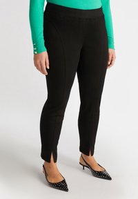 Pantalones negros ajustados con aberturas en los tobillos, hechos de tela elástica. Llevados con tacones de punta abiertas negros con tachuelas, combinados con una blusa verde.