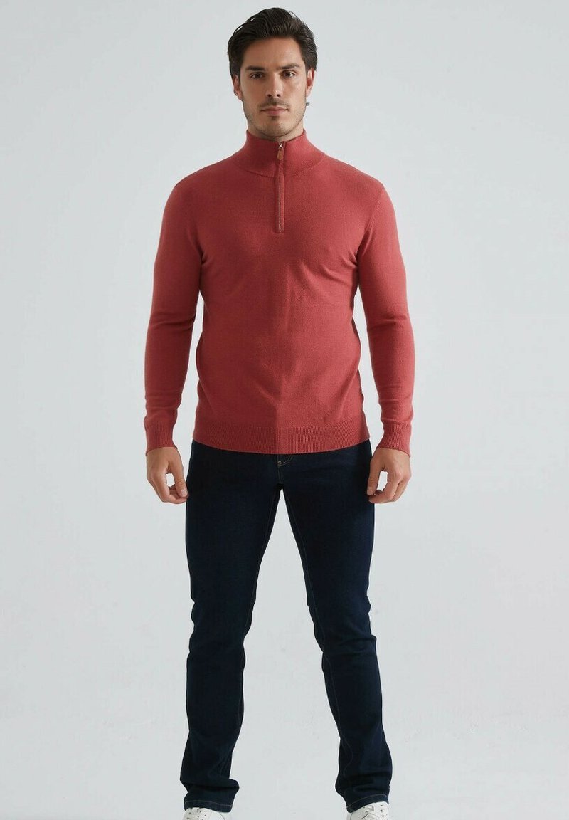 Sweatshirt rouge à manches longues avec zip, au design ajusté, avec des poignets côtelés et une texture lisse, associé à un jean bleu foncé et des baskets blanches.