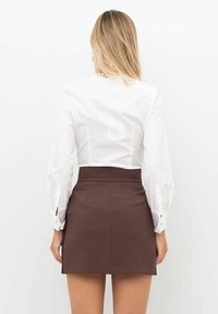 Camicia bianca a maniche lunghe con maniche a sbuffo, abbinata a una gonna marrone a vita alta. Presenta una vestibilità sagomata e bottoni decorativi ai polsini.