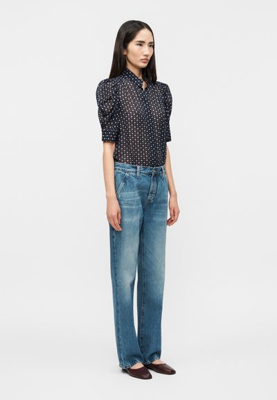 Γυναίκα με μακριά σκούρα μαλλιά, που φοράει διάφανη μαύρη μπλούζα με άσπρες βούλες, μανίκια με όγκο, φαρδιά μπλε τζιν και σκοτεινές μυτερές μπαλαρίνες, στέκεται πλευρικά.