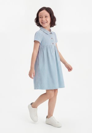 Χαμογελαστή νεαρή κοπέλα που φοράει φόρεμα με γαλάζιες και λευκές ρίγες και κουμπιά, περπατάει με λευκά αθλητικά σε ένα λευκό, απλό φόντο.