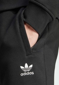 Svarta bomullsjeans med sidoficka, med en vit Adidas trefoil-logotyp. Mjuk textur, elastiskt midjeband och minimalistisk design.