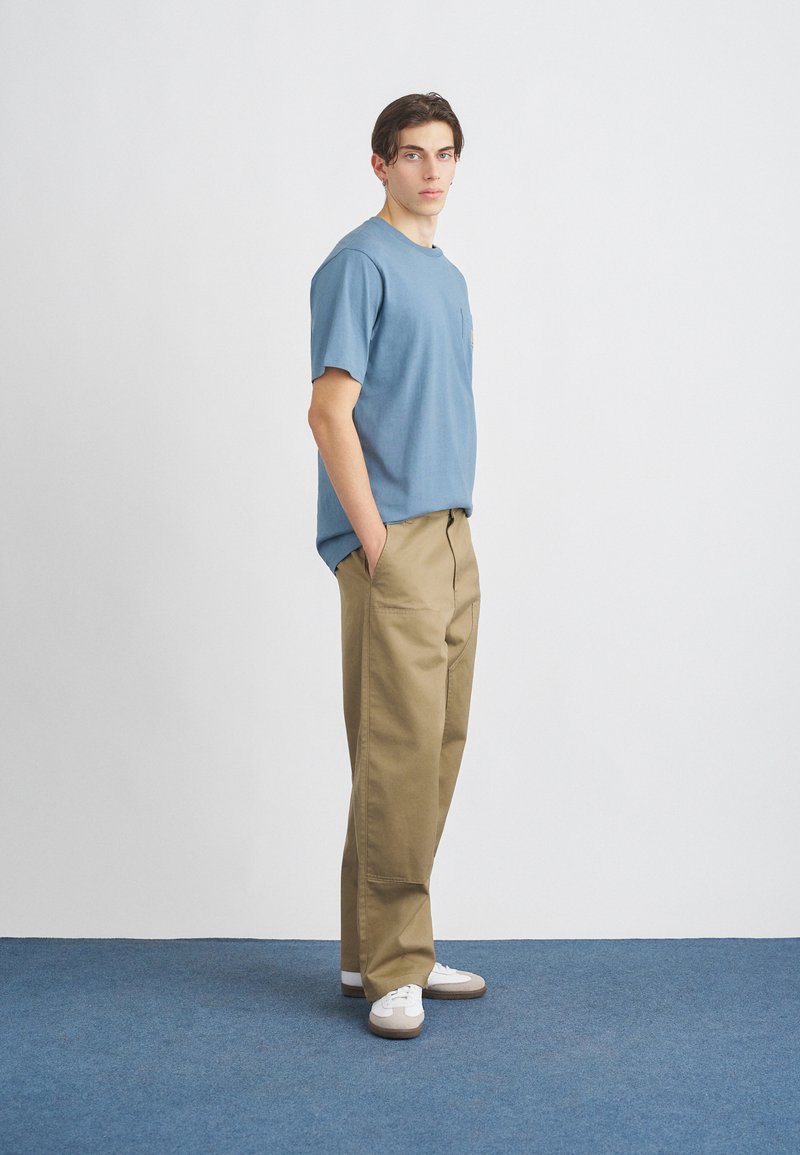 Ljusblå bomulls-T-shirt med bröstficka, tillsammans med sandfärgade högmidjade byxor. Vita sneakers fulländar outfiten.