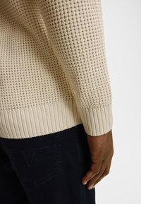 Beżowy sweter z dzianiny o teksturowanym splocie i ściągaczach na rękawach, zestawiony z granatowymi jeansami z przeszyciem na kieszeni.