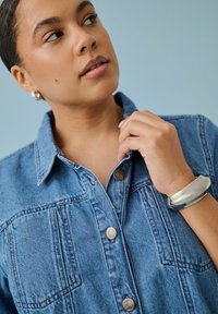 Femme portant une veste en jean bleue, tenant le col avec sa main droite, portant des boucles d'oreilles créoles argentées et un gros bracelet en argent.