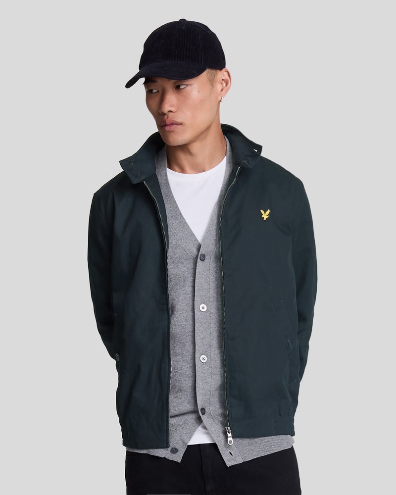 Marineblaues Cordhut, dunkelgrüne Jacke mit Kragen und Reißverschlüssen, graue Strickjacke mit Knöpfen, weiße Bluse darunter, schwarze Hose.