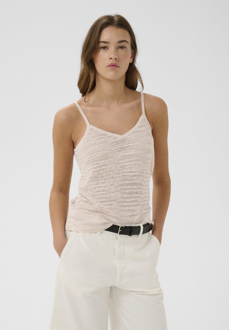 Jeune femme aux cheveux bruns portant un haut beige sans manches texturé et un pantalon blanc taille haute avec une ceinture noire, les mains dans les poches.