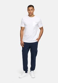 Weißes T-Shirt mit Rundhalsausschnitt und Brusttasche, kombiniert mit navyblauen Cargo-Hosen mit Seitentaschen und elastischen Bündchen, ergänzt durch weiße Sneakers.
