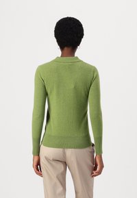 Osoba z krótkimi kręconymi włosami, ubrana w dopasowany zielony sweter z długim rękawem i beżowe spodnie, stojąca na tle gładkiej białej ściany.