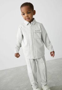 Harmaa vetoketjullinen takki, jossa on teksturoitu kuvio ja sivutasku, yhdistettynä harmaihin housuihin, joissa on hienovaraiset sauman yksityiskohdat.