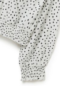 Witte stof met zwarte hartpatronen; heeft gathered mouwen voor een aansluitende vorm. Glad textuur, lichtgewicht materiaal.