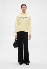 Žltá mikina s kapucňou s nápisom "GANT1949", kombinovaná s čiernymi širokými nohavicami a malou čiernou taškou. Hladké, príležitostné textúry látok.