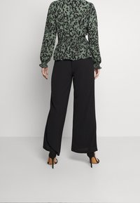 Bluse mit grünem und schwarzem Tiermuster, elastischem Bund, kombiniert mit lockeren schwarzen Hosen und schwarzen High-Heels.