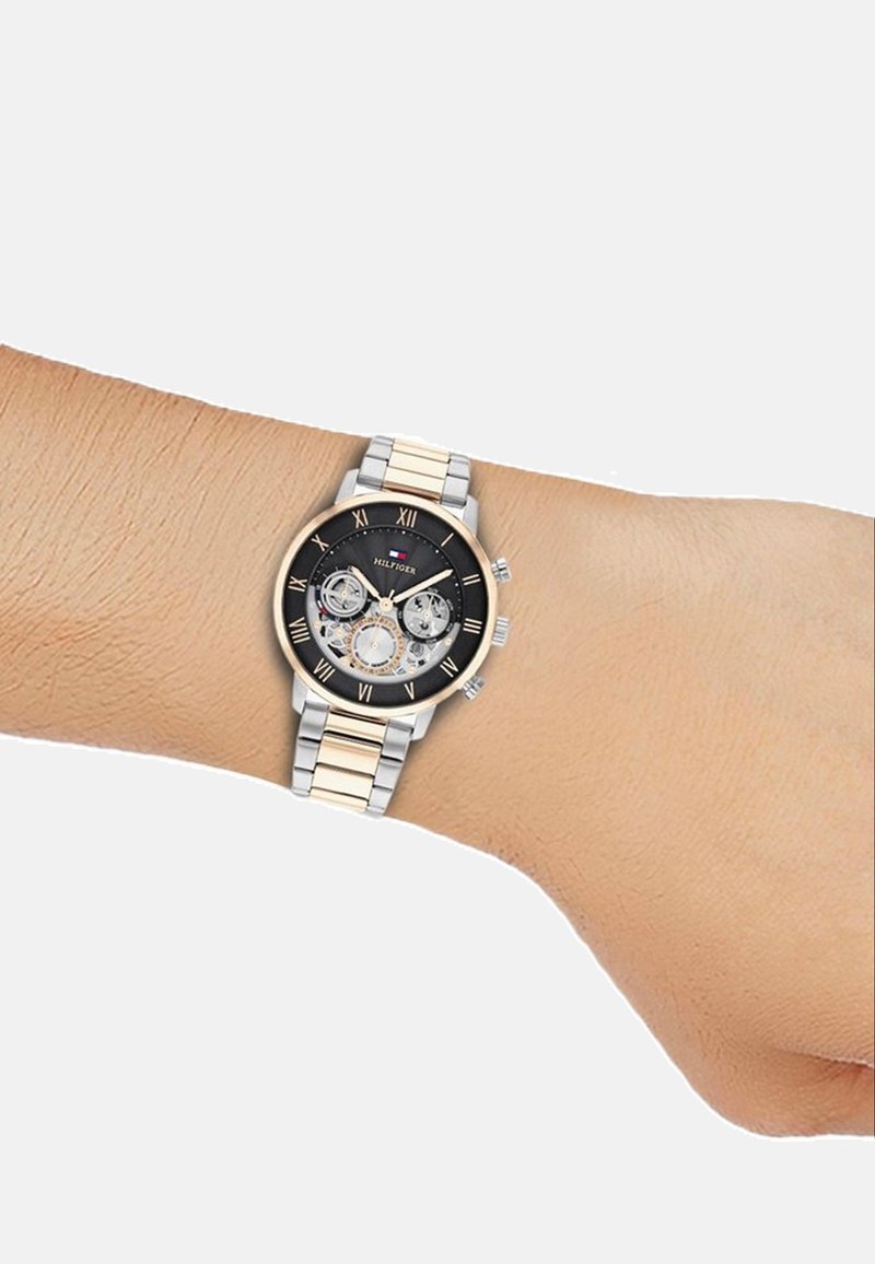 Svart kronografklocka med tre underdials, silver- och guldfärgad metallarmband, rund urtavla, romerska siffror som markörer och en framträdande logotyp.