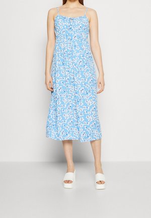 Vrouw draagt een lichtblauwe midi-jurk met witte bloemenprint en dunne bandjes, gecombineerd met witte open sandalen met plateauzool.