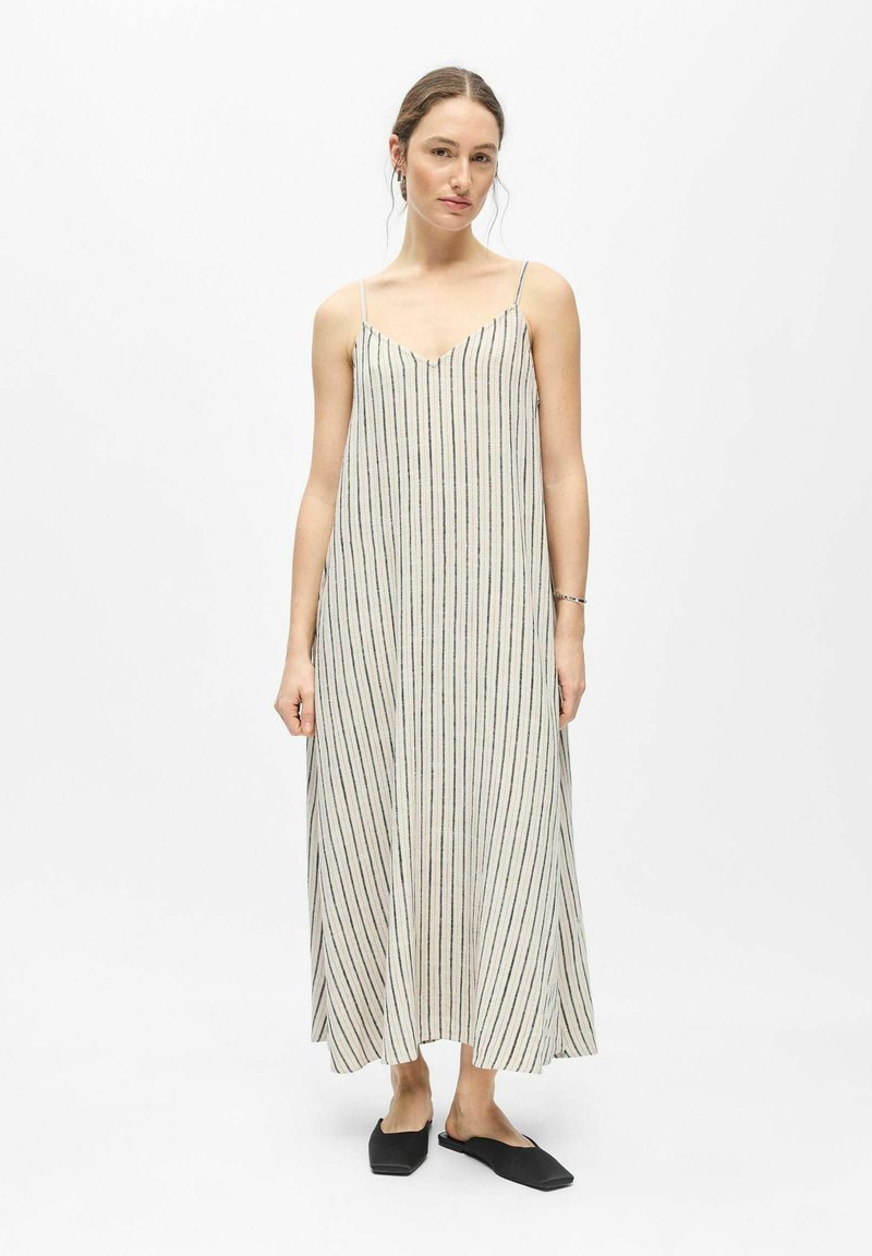 Femme debout, portant une longue robe rayée sans manches avec de fines bretelles et des chaussures noires sans lacets, sur un fond blanc uni.