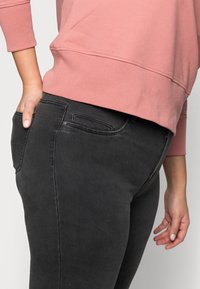 Black denim jeans with a slim fit, featuring a back pocket, and paired with a light pink long-sleeve sweatshirt. The fabric appears soft.