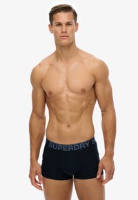Czarne bokserki z kontrastującym szarym pasem na biodrach z napisem "SUPERDRY". Materiał wydaje się gładki i dopasowany. Model jest bez koszulki.