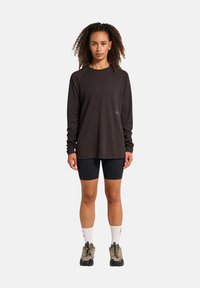 Frau mit lockigem Haar, die ein schwarzes Langarmshirt, schwarze Radlerhosen, weiße Socken und Wanderschuhe trägt, steht vor einem weißen Hintergrund.