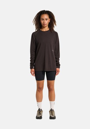 Vrouw met krullend haar, gekleed in een zwart longsleeve shirt, zwarte fietsbroek, witte sokken en wandelschoenen, staand op een witte achtergrond.