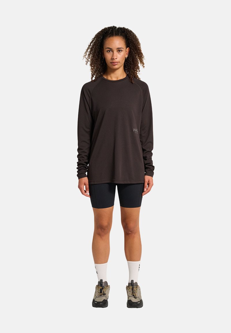 Frau mit lockigem Haar, die ein schwarzes Langarmshirt, schwarze Radlerhosen, weiße Socken und Wanderschuhe trägt, steht vor einem weißen Hintergrund.