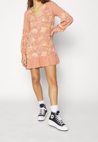 Kvinde iført en ferskenfarvet blomster mini kjole med flæser i nederdelen og sorte høj-top Converse sneakers med hvide sokker.