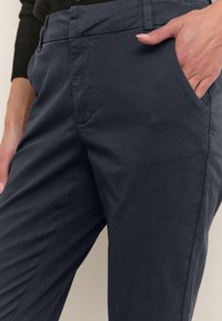 Tmavě šedé bavlněné kalhoty slim fit, s bočními kapsami a viditelnými švy, zobrazené s rukou spočívající v kapse.