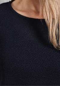 Mörkblått stickat tröja med rund halsringning. Strukturen i tyget är synlig, vilket framhäver ett löst stickat mönster. Slät, mjuk utseende.