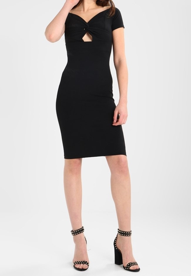 Femme portant une robe noire près du corps longueur genou avec une découpe devant et des manches courtes, associée à des sandales noires à talons hauts cloutées.