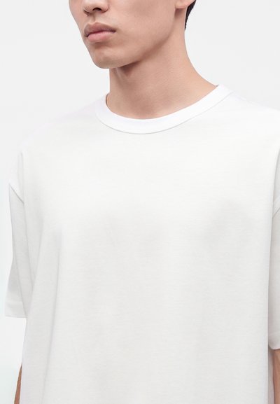 Gros plan d'une personne portant un t-shirt blanc uni à col rond et manches courtes sur un fond clair.
