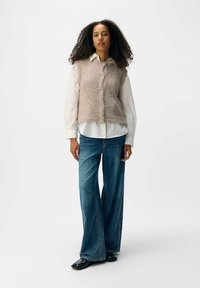 Hellbrauner gestrickter Westen mit strukturiertem Finish, Knopfleiste vorne, über einem weißen Hemd getragen, kombiniert mit weit geschnittenen blauen Jeans und schwarzen Schuhen.